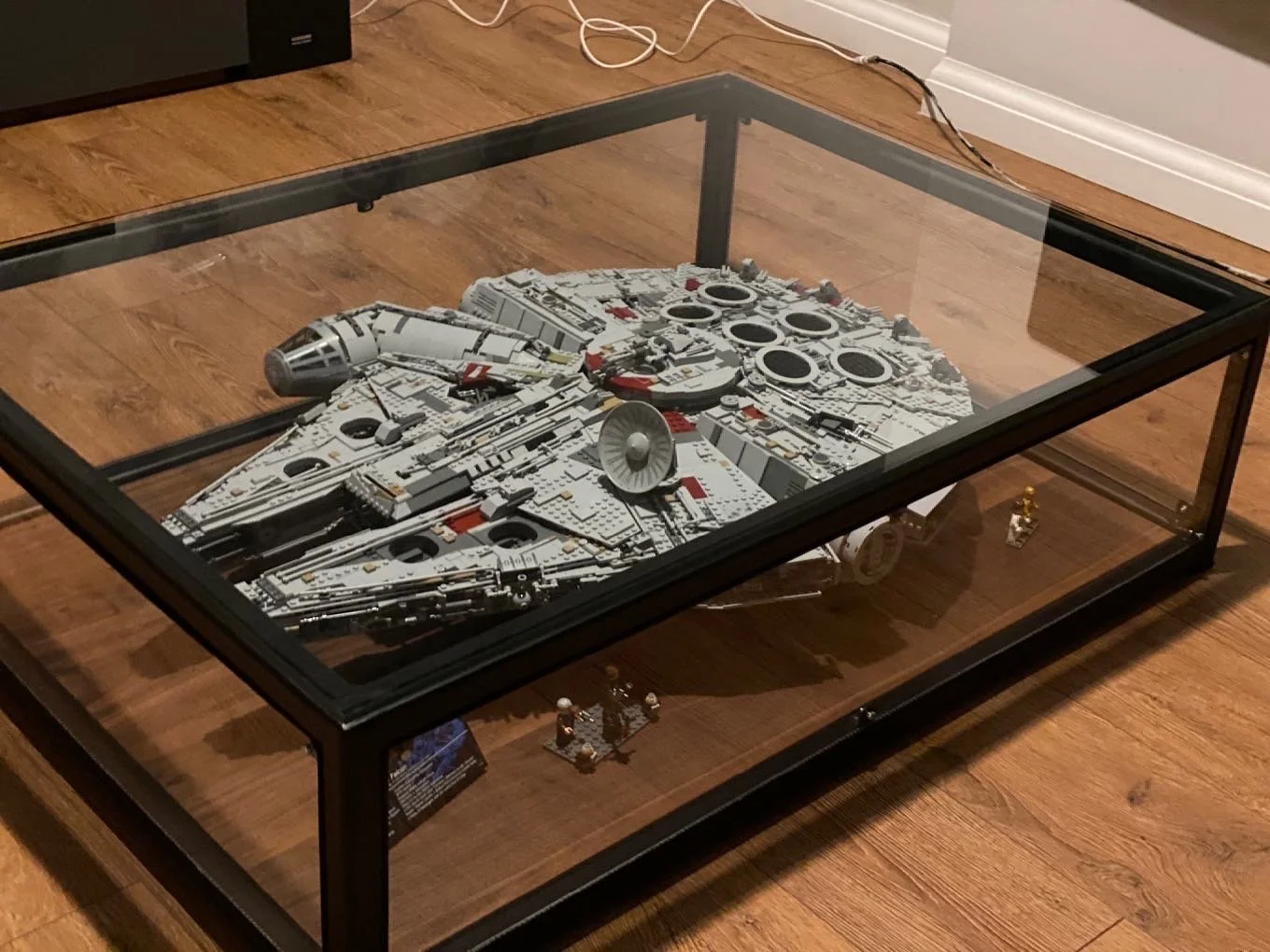 Glass‑top coffee table with a black metal frame displaying a LEGO Millennium Falcon model inside.