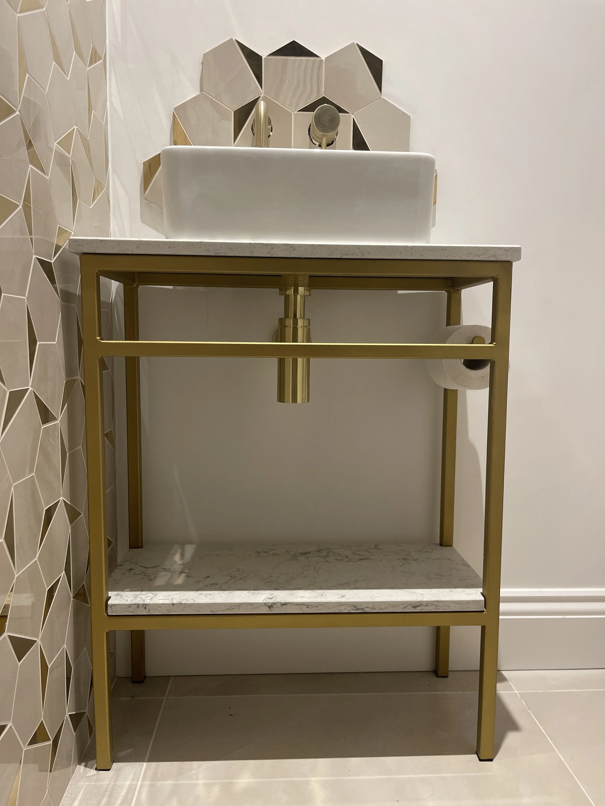Modern bathroom vanity with a gold metal frame, marble panels, white rectangular sink, and geometric mirror tiles.