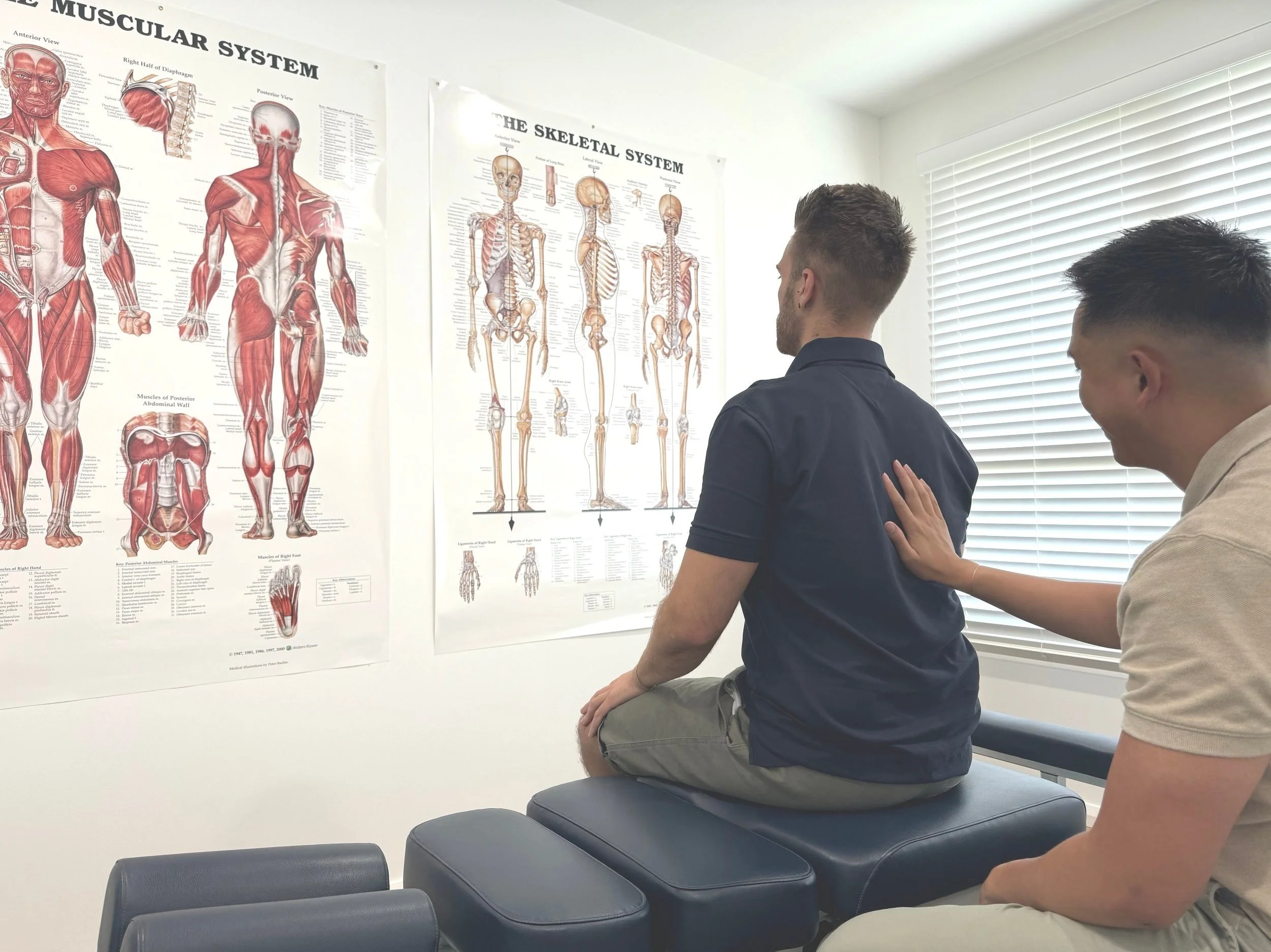 A man receives physical therapy on his shoulder from a healthcare provider in a room with anatomical posters of the muscular and skeletal systems.