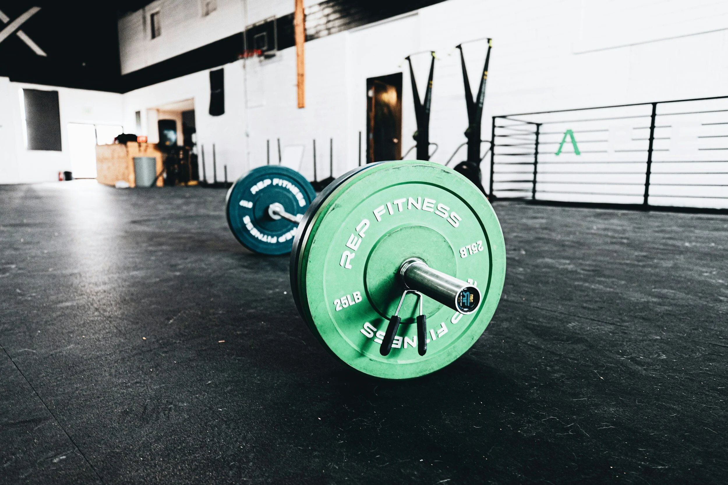Barbell with green weight plates on a black gym floor, with gym equipment and white walls in the background.