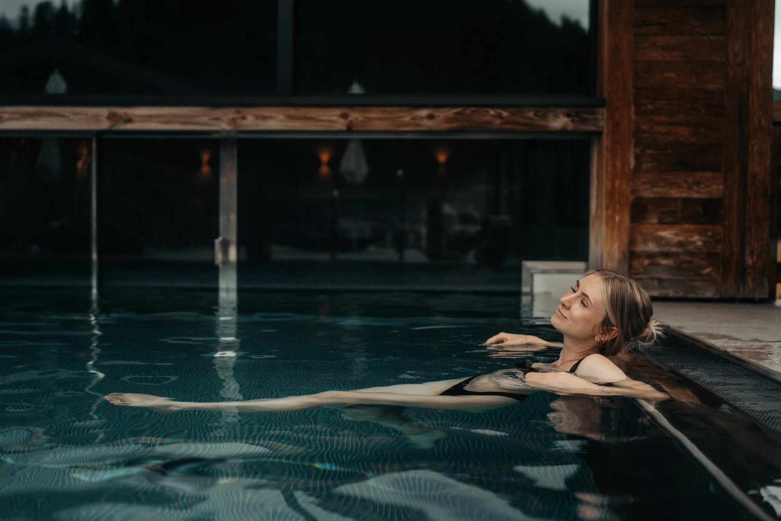 Eine junge Frau entspannt in einem Pool, lehnt an der Poolkante, mit Holzhintergrund im Hintergrund.