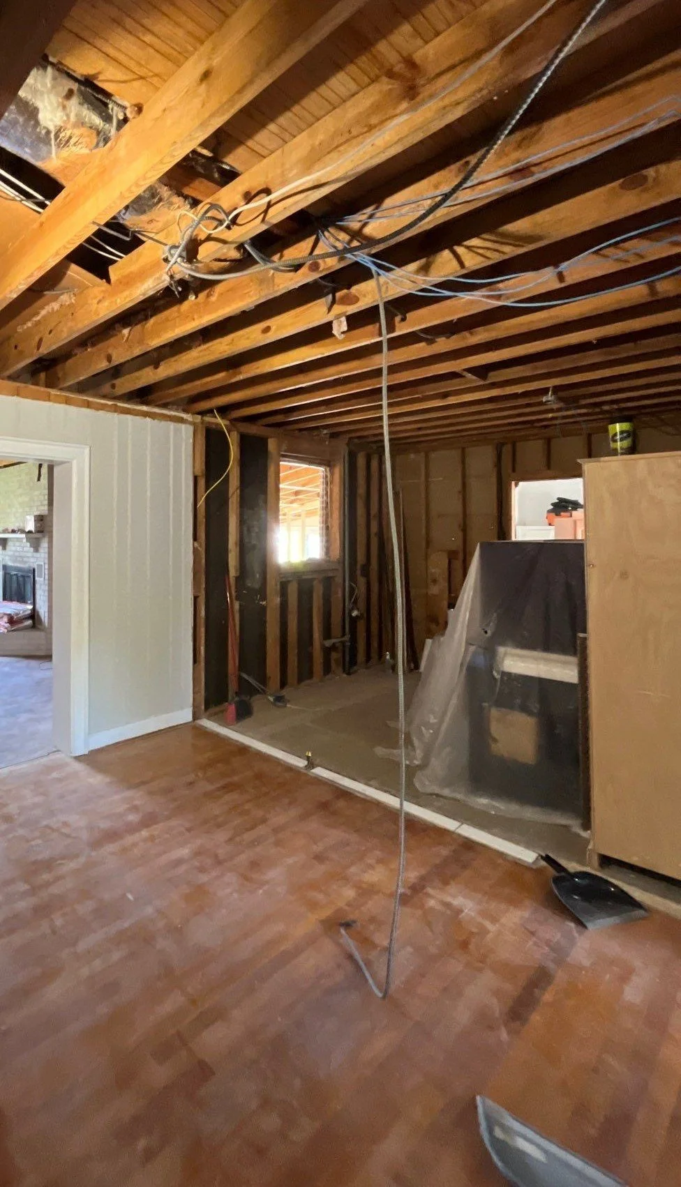 Interior of a house under renovation, showing exposed ceiling beams with electrical wires, partially framed walls, a doorway opening to another room, and construction materials and tools scattered around.