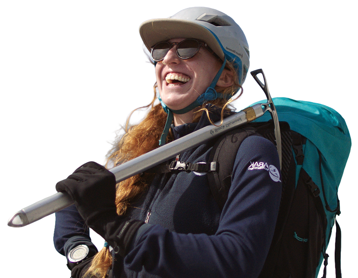 An Alaskan guide with long red hair, wearing outdoor gear including a helmet, sunglasses, and a backpack, is smiling happily while holding a pole.
