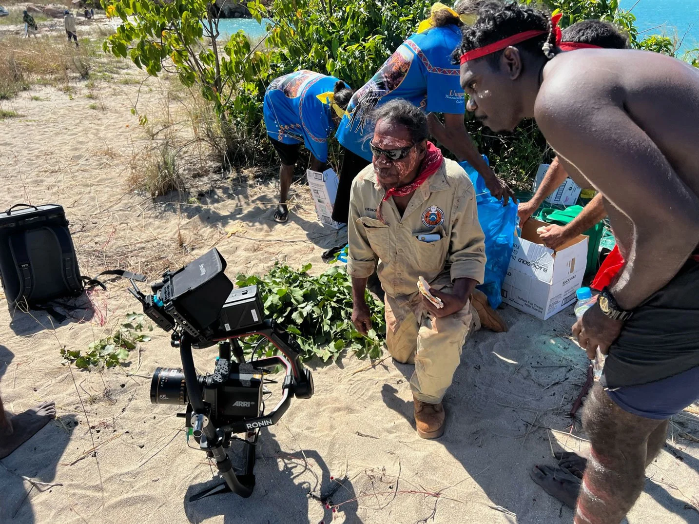 Kimberley Australia Cruise. Group of people, including a man kneeling in beige uniform, gathered around a small plant on a sandy beach, with photography equipment and boxes nearby, and ocean in the background.