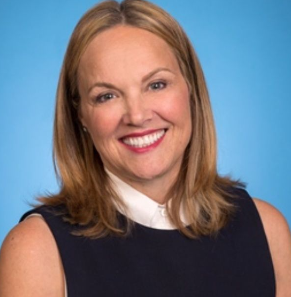 Smiling woman with wavy blonde hair, wearing a sleeveless navy dress with a white collar, against a blue background.