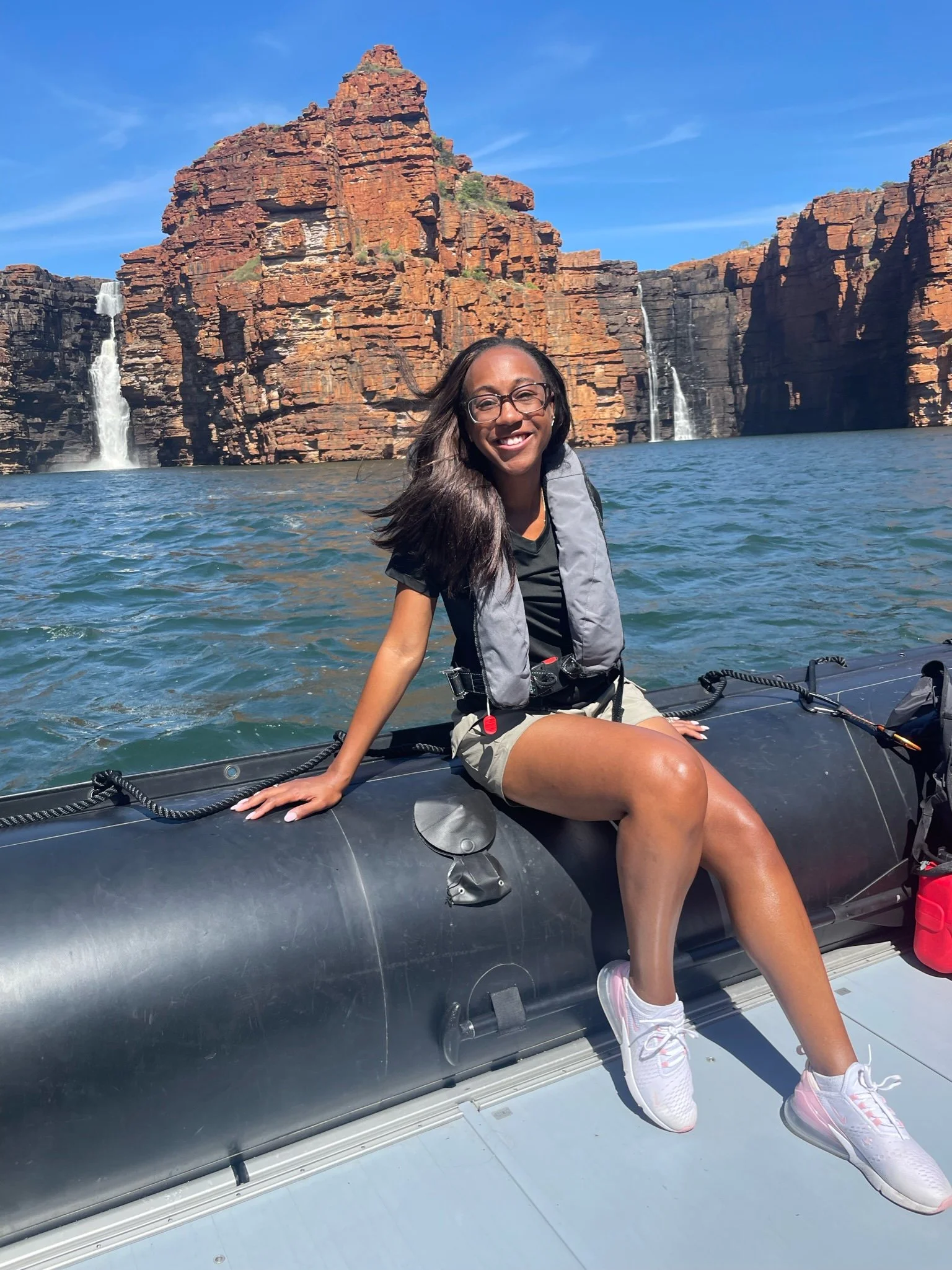 A woman sitting on the edge of a boat with water, red rock formations, and waterfalls in the background in Kimberley Australia. 