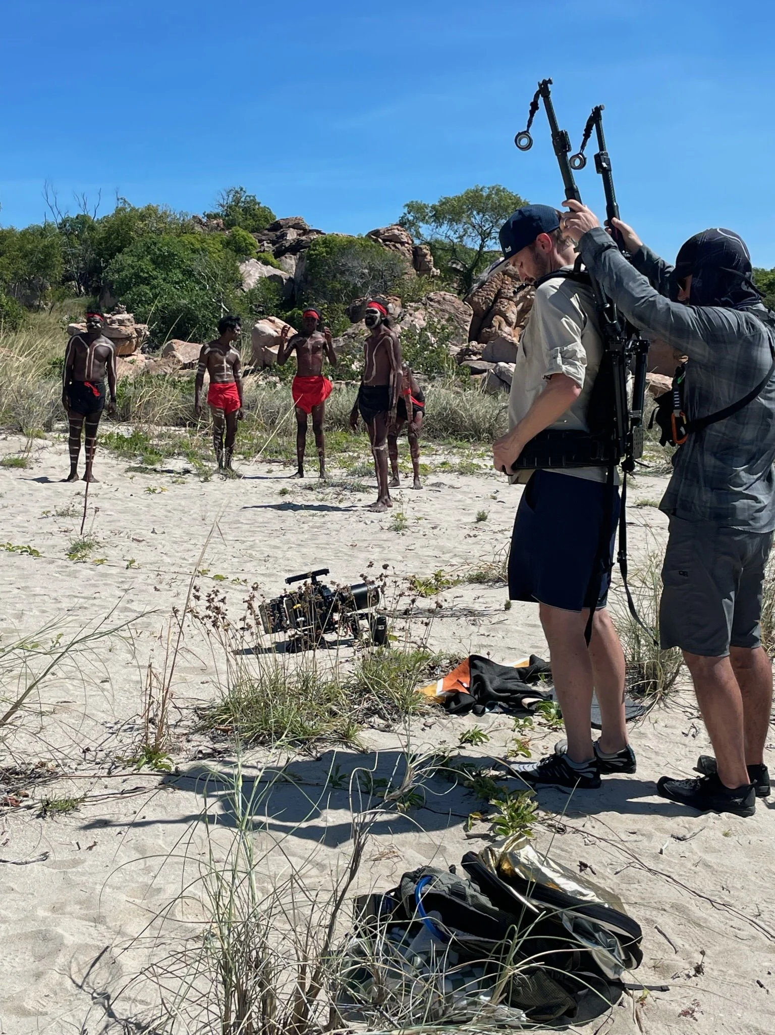 Filming crew setting up a scene with indigenous people in traditional attire and face paint at a beach with rocks and greenery, under a clear blue sky in Kimberley Australia. 