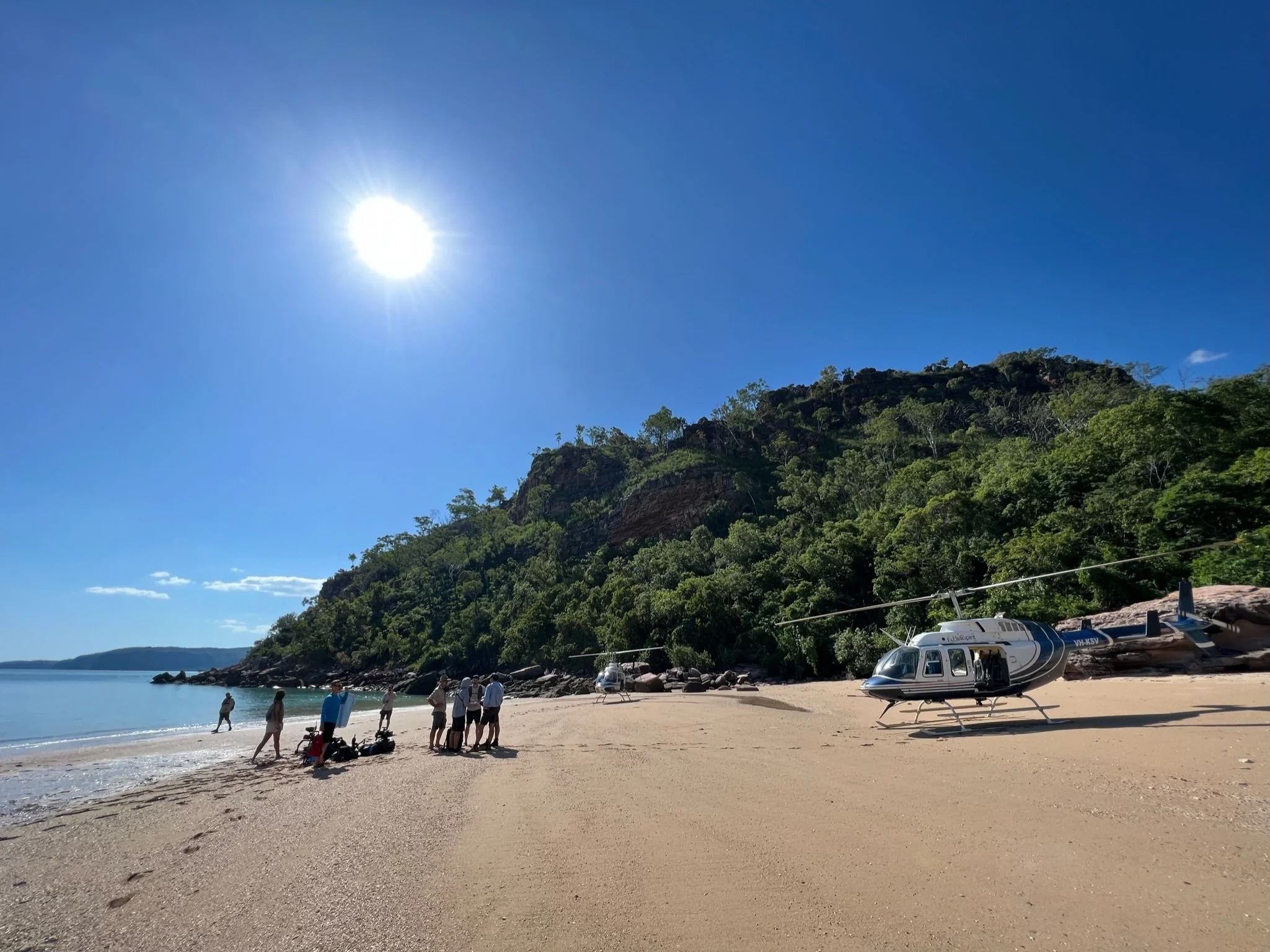 A sandy beach with a helicopter parked on the sand near a group of people. A lush green hill rises behind the beach under a bright blue sky with the sun shining in Kimberley Australia. 