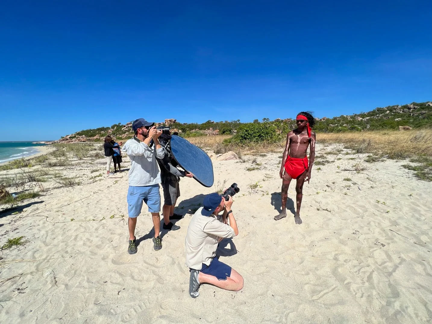 Kimberley Australia Cruise. A photoshoot happening on a sandy beach with a man dressed as a Native American in red loincloth and headband, photographers capturing him with professional gear, and some people observing in the background.