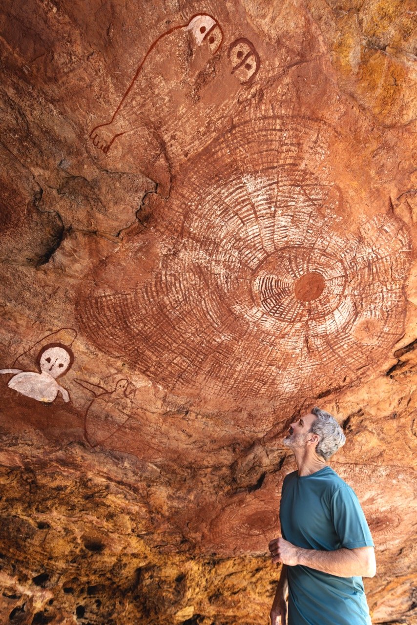 Kimberley Australia Cruise. A man with gray hair in a teal shirt looking up at cave paintings and engravings on a rocky ceiling, including stick figures and a large circular pattern.