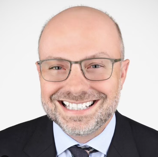 Portrait of a man with glasses smiling in a suit and tie against a white background.