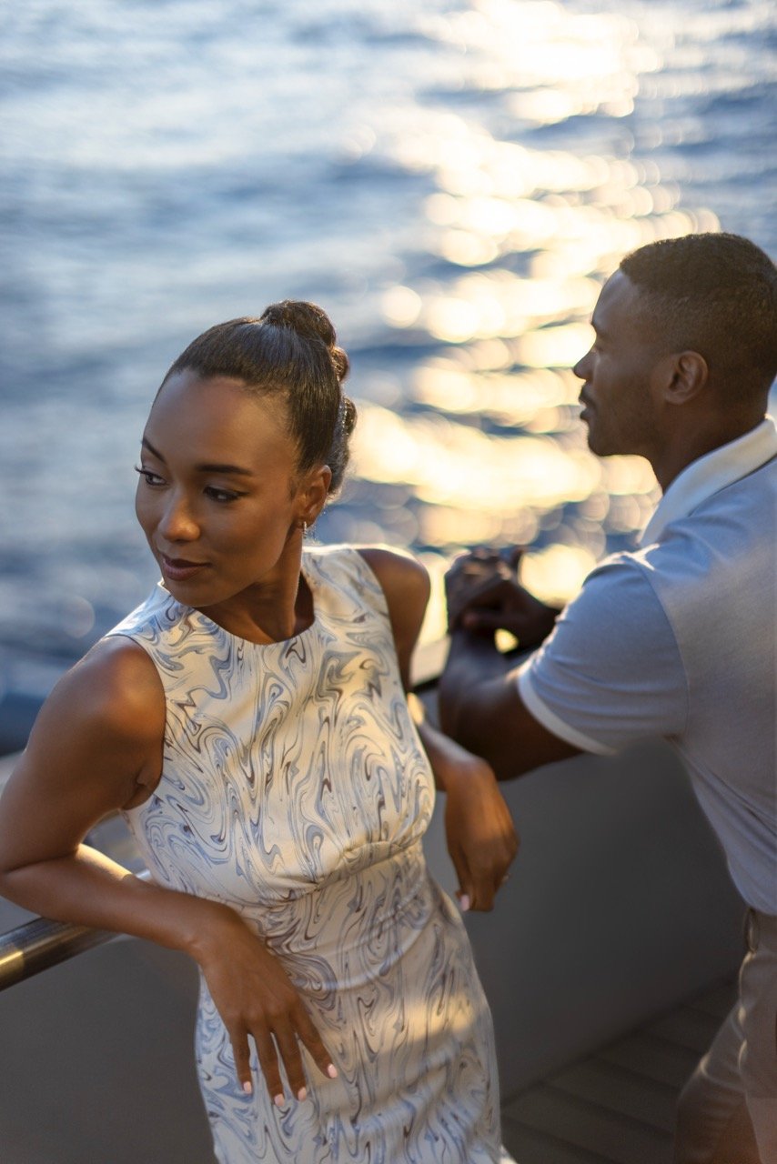 A woman in a marble-patterned sleeveless dress stands on a boat, leaning against the railing, with a man in a light-colored polo shirt and shorts beside her, both looking toward the water during sunset.
