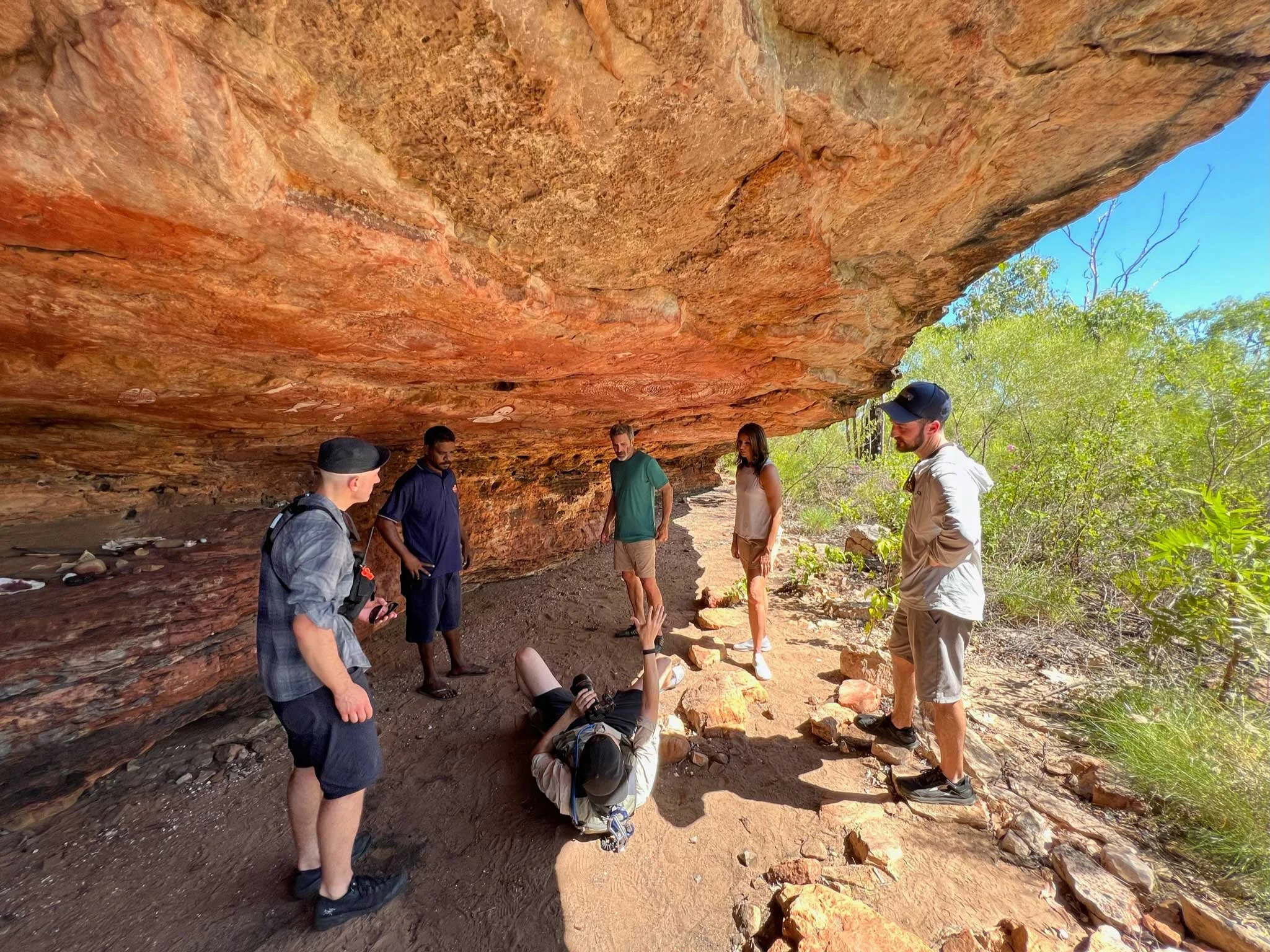 Group of people gathered under a large orange and brown rock overhang on a trail, with a woman lying on the ground gesturing and four others standing around, surrounded by green vegetation and clear blue sky.