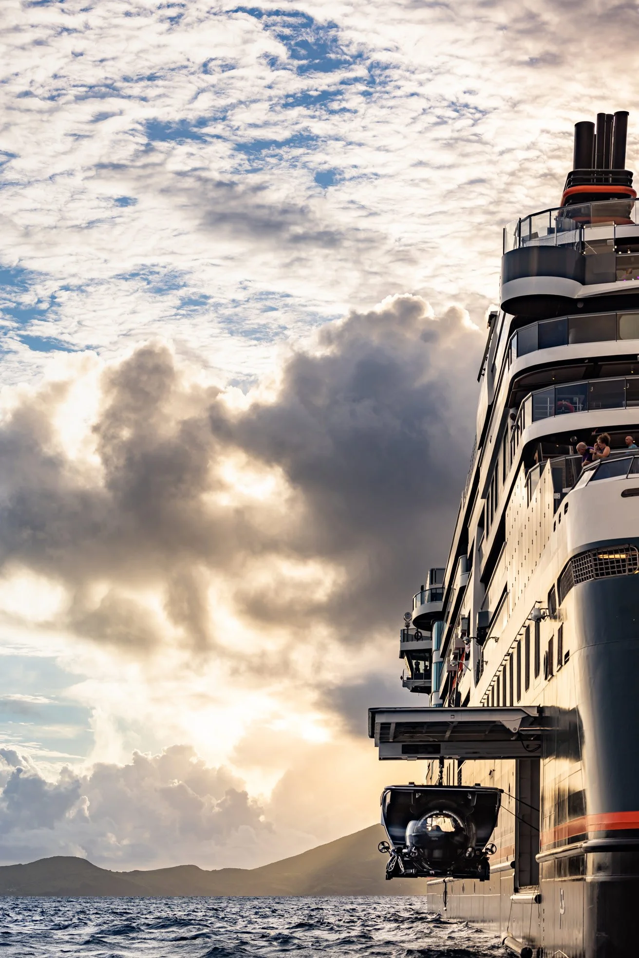 A large cruise ship sailing on the ocean during a sunset with mountains in the background and a partly cloudy sky overhead.