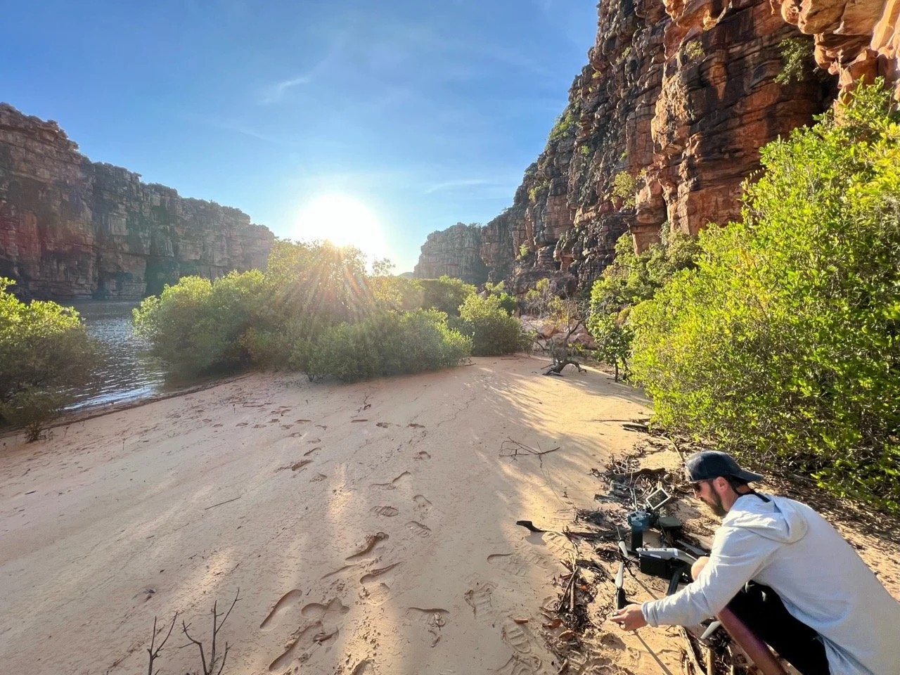 Kimberley Australia Cruise. A person with a beard, sunglasses, and cap kneeling on sandy terrain assembling or adjusting camera equipment, with footprints in the sand, green bushes, rocky cliffs, and a river at sunset in the background.