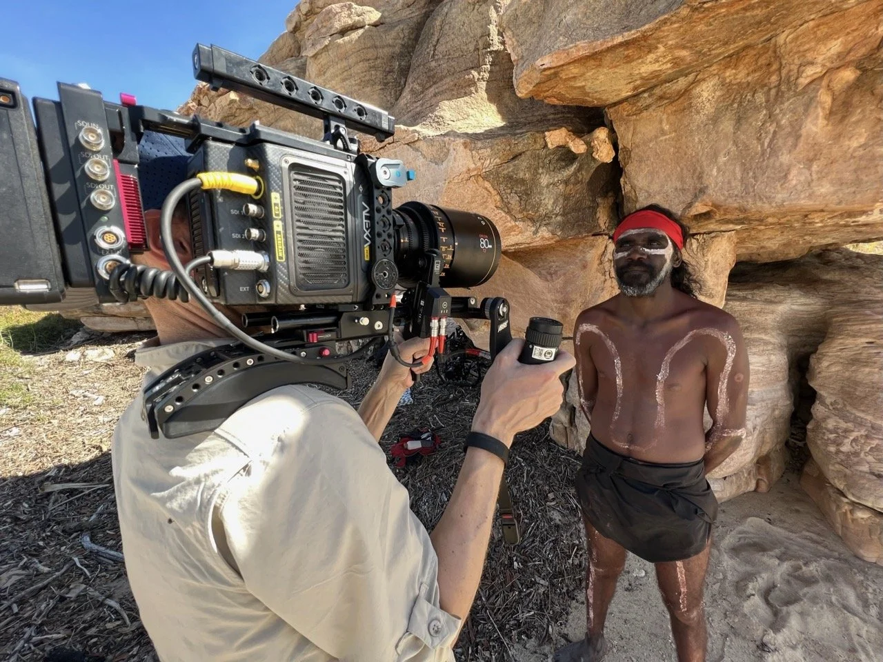 Kimberley Australia Cruise. Filmmaker operating camera outdoors with man in traditional attire, pink markings on body, red headband, sunglasses, sandy ground and large rocks in background.