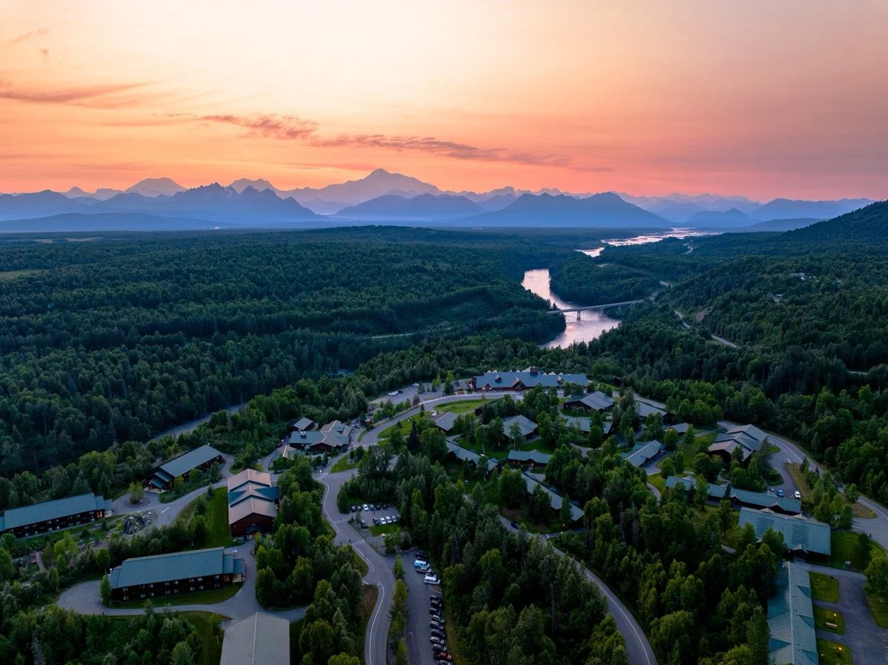 Alaska Cruise. Aerial view of a green forested landscape with a small neighborhood of buildings and roads in the foreground, a river winding through the hills, and mountains in the distance at sunset with pink and orange sky.