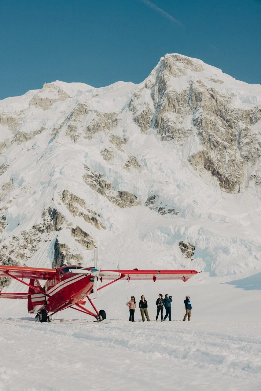 A red airplane parked on snow with a group of five people standing nearby, mountains and glaciers in the background under a clear blue sky.