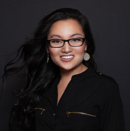 A woman with long dark hair, glasses, and earrings, smiling against a dark background.
