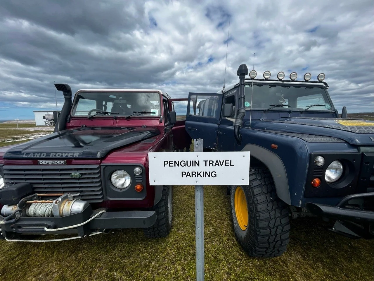 Two off-road vehicles, a red Land Rover Defender and a blue one with additional lights on the roof, parked side by side in a grassy area under a cloudy sky, with a sign reading "Penguin Travel Parking" in front of them.