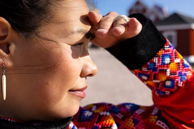A woman with earrings uses her hand to shield her eyes from the sun while outdoors, wearing a colorful beaded garment or accessory.
