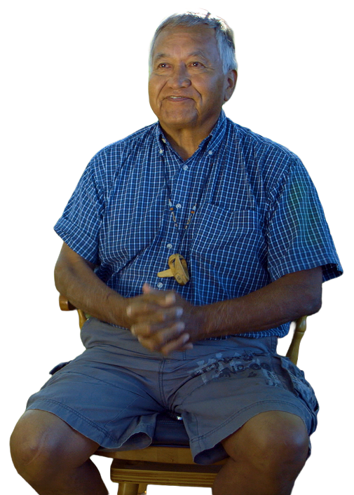 An elderly Alaskan native man sitting on a wooden chair, wearing a blue checkered short-sleeve shirt and gray shorts, with glasses hanging around his neck, smiling and clasping his hands together.