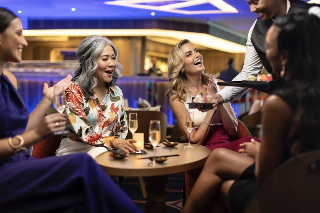 A group of five women socializing and laughing at a restaurant or bar, with drinks on the table, while a waiter serves desserts.