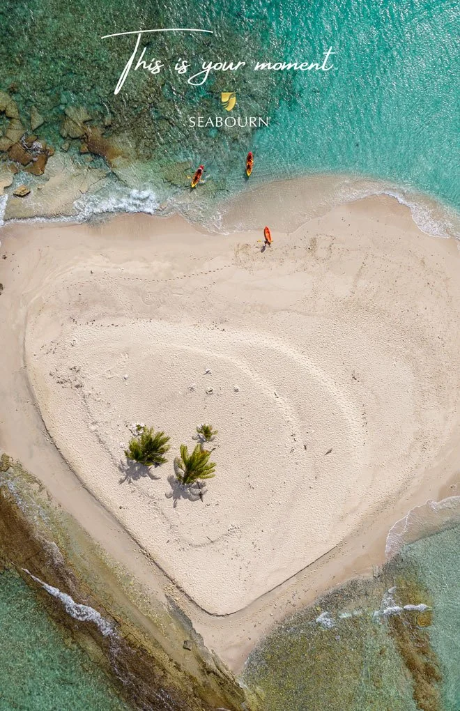 A heart-shaped sandy beach surrounded by turquoise water with a few palm trees in the center, and three people with kayaks near the shore. Text overlay says 'This is your moment' and 'SEABOURN'. WABU Creative, Scott David Martin