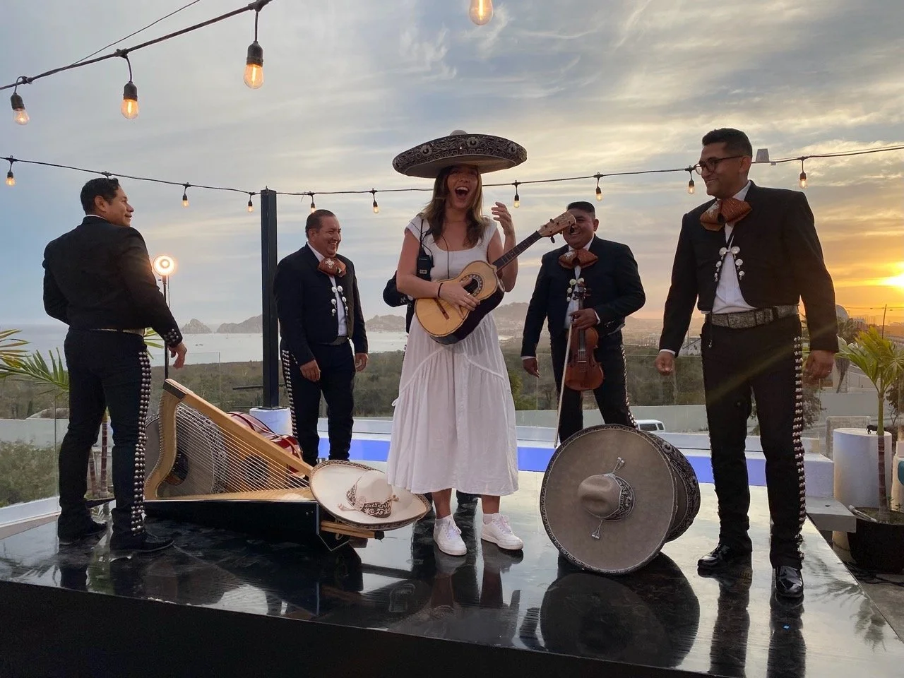 Behind the Scenes of WABU Creative. Group of musicians performing outdoors during sunset, with women in traditional Mexicano attire and men in charro outfits, and Producer Destiny playing guitar, in a setting with string lights and ocean view.