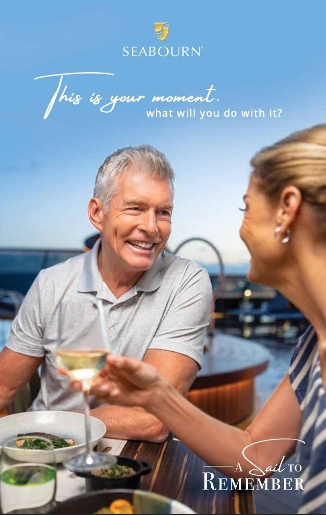 An older man and woman enjoying a meal together at a restaurant near the water, smiling at each other. The man is holding a glass of white wine. The image is part of a Seabourn advertisement with the text 'This is your moment. What will you do with i