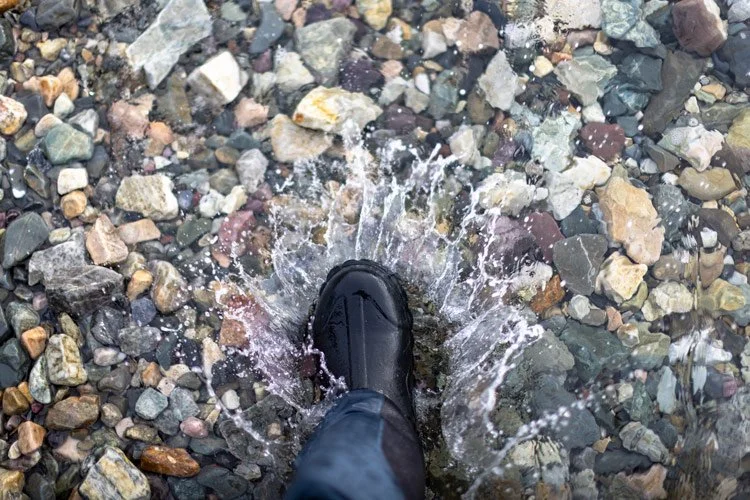 A person wearing a black boot stepping into a shallow puddle on a bed of multicolored rocks.