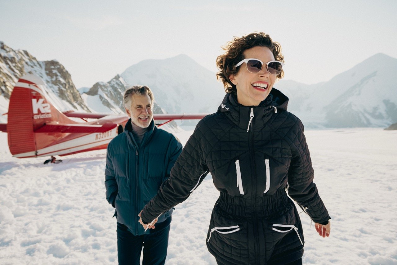 Alaska Cruise. A woman in sunglasses and a black winter jacket, smiling, in a snowy mountain landscape. A man in a blue jacket is smiling behind her. A red small airplane is on the snow near the mountains.