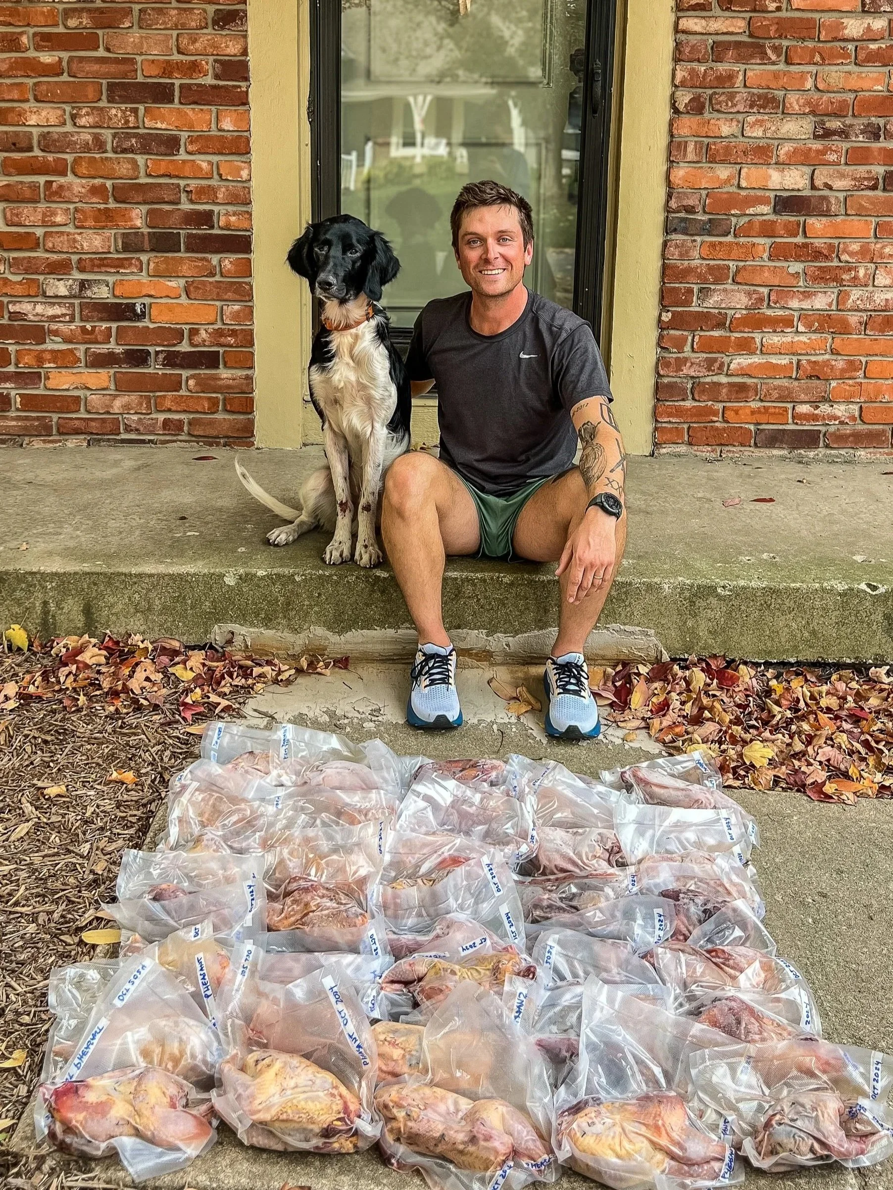 A man sitting on the porch with a black and white dog, surrounded by bags of raw chicken breasts laid out on the ground in front of them, in front of a brick house with the number 4000 on the wall.