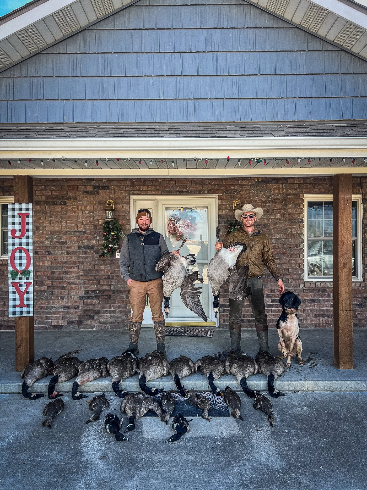 Two men stand on a porch with a brick house in the background, holding ducks they hunted. There are more ducks laid out on the ground in front of them, and a dog sitting to the right. A decorative 