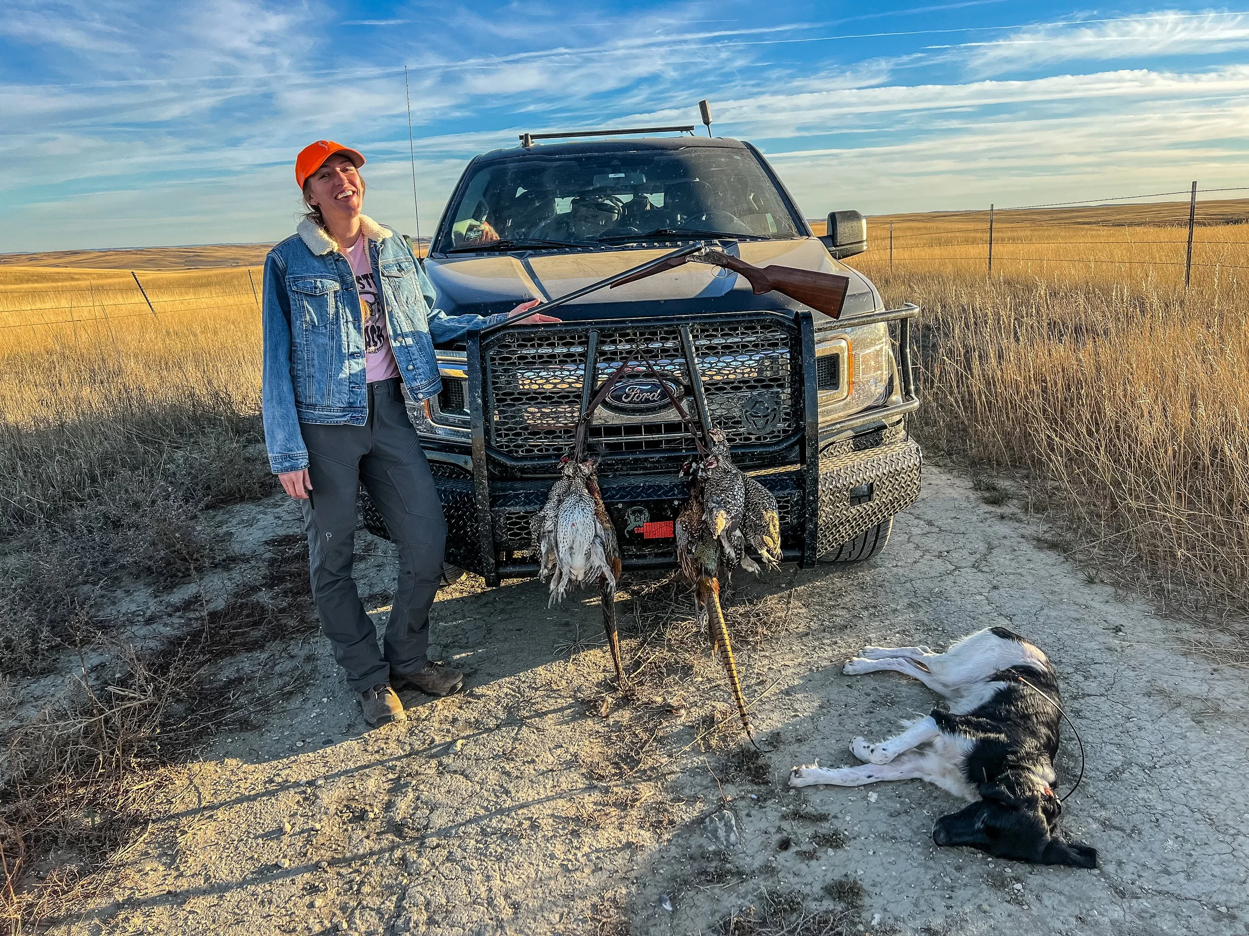 A woman standing next to a pickup truck with a front grille guard, holding a hunting rifle. She is smiling and wearing a denim jacket and an orange cap. In front of the truck on the ground, there are two dead ducks hanging from a rack, a dog lying do