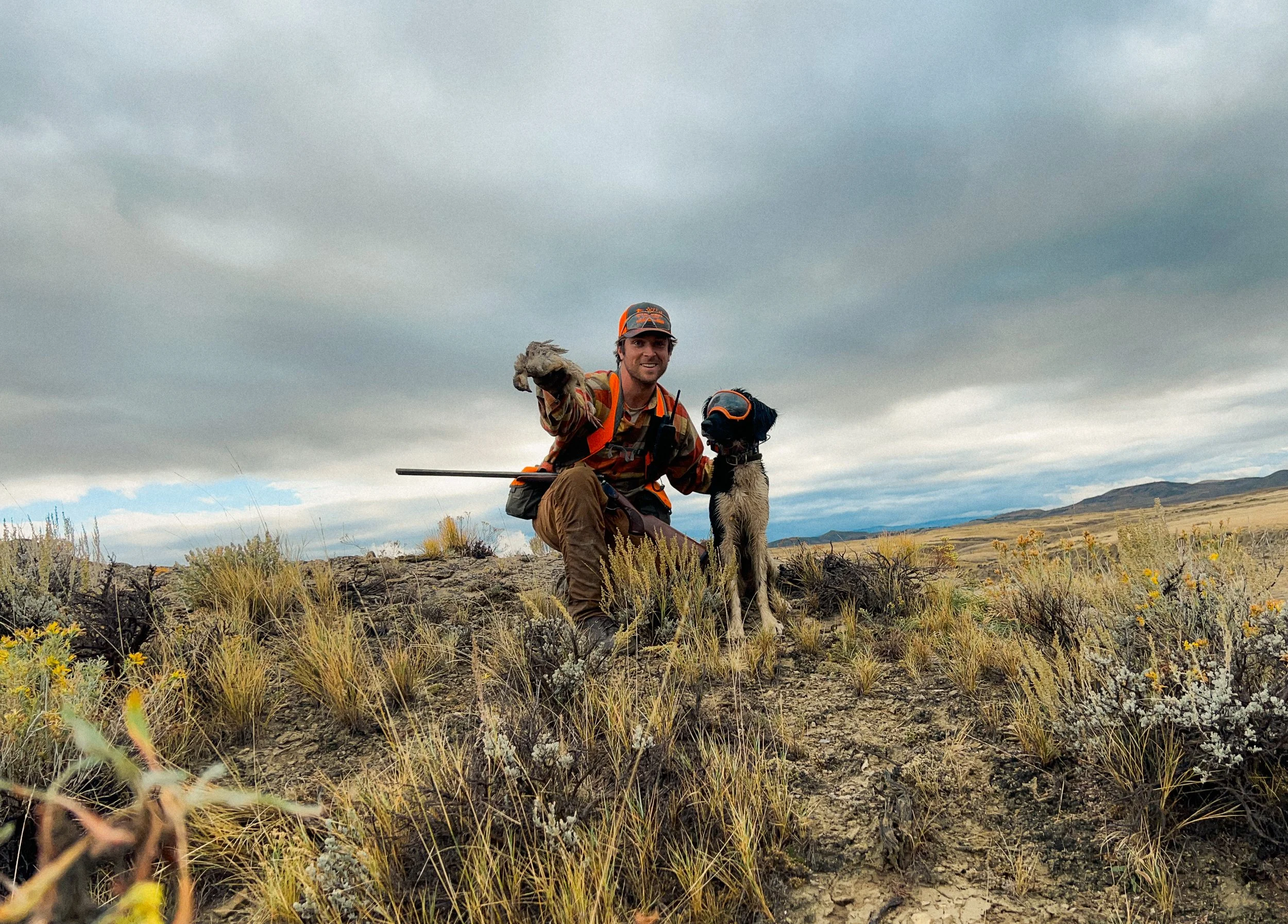 A man in outdoor gear kneeling on rocky, grassy terrain, pointing, with a dog wearing an orange vest and goggles next to him, under a cloudy sky with mountains in the distance. Chukar partridge, Wyoming.