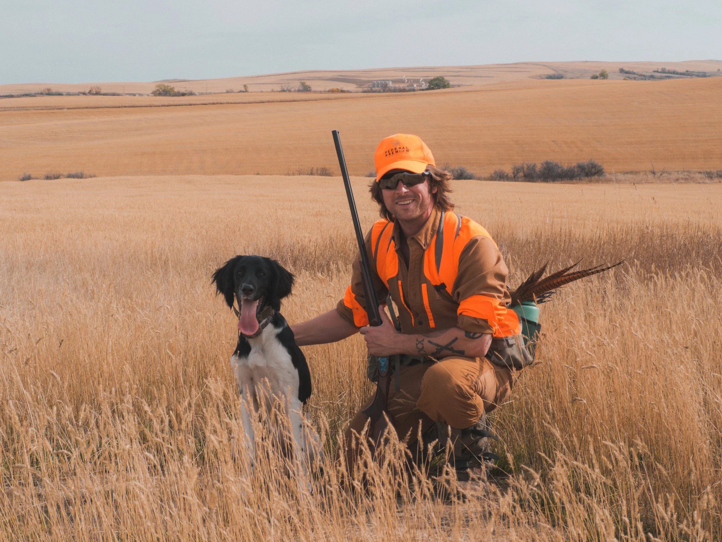 A man in orange and brown camouflage hunting gear crouching in a grassy field with a black and white dog, holding a shotgun, in a rural landscape with rolling hills.