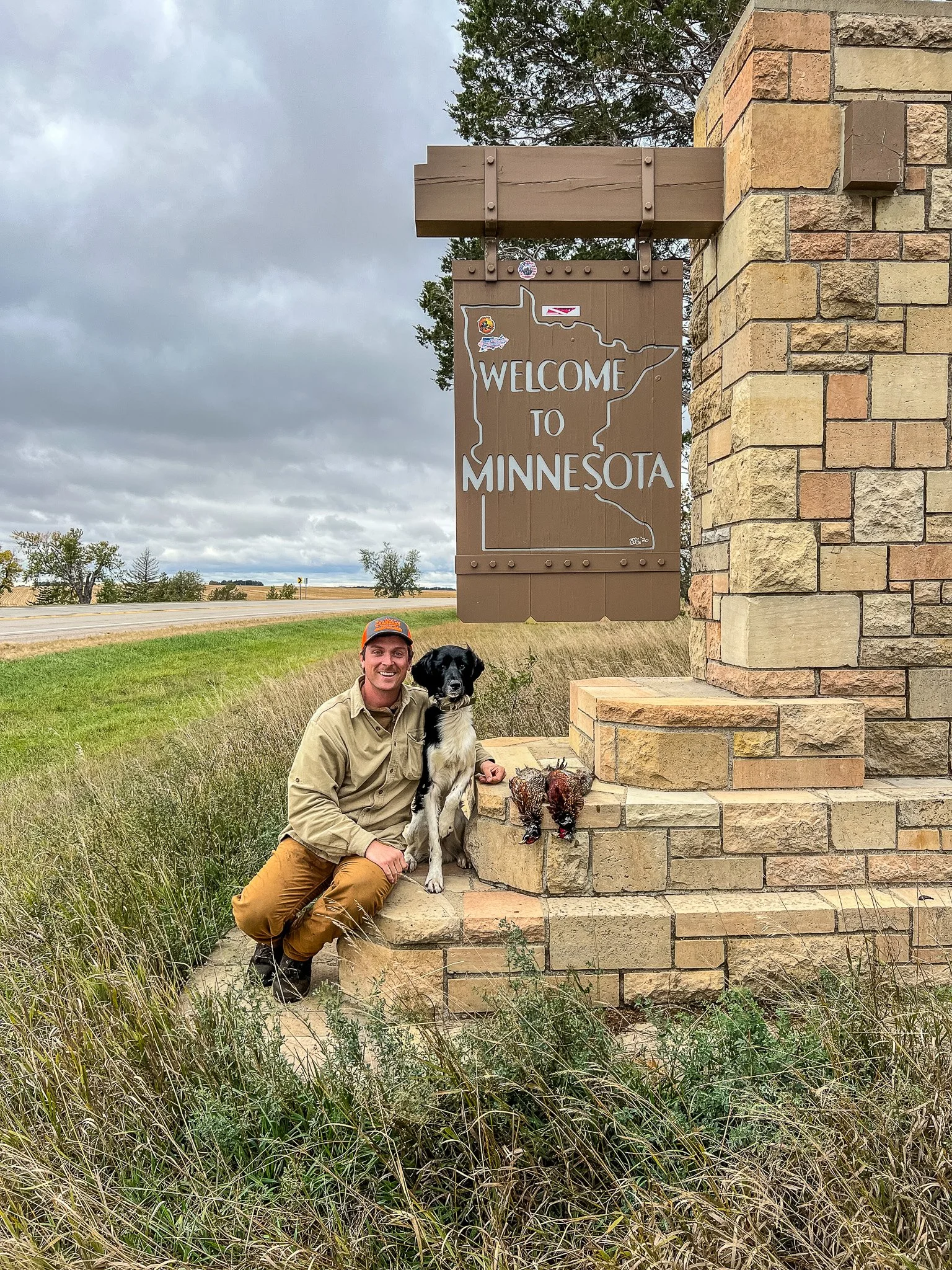 Aldo, a Large Munsterlander versatile hunting dog, with Ian Burrow, posing with their daily limit of Ring-neck rooster pheasants beside the Welcome to Minnesota sign after pheasant season opening day in Minnesota.