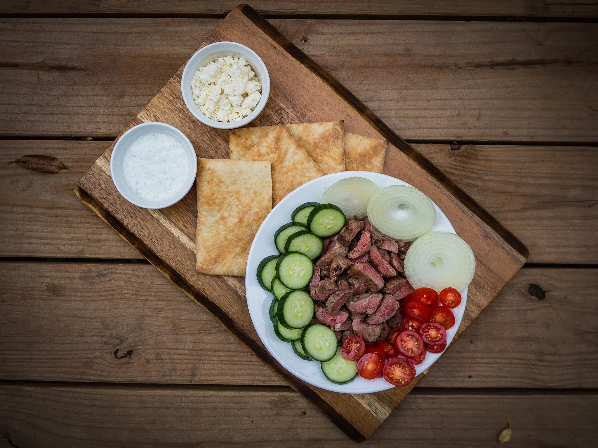 A wooden board with a white plate of sliced cucumbers, cherry tomatoes, sliced onions, and cooked steak, along with two small bowls of crumbled cheese and creamy dressing, and two pieces of flatbread.
