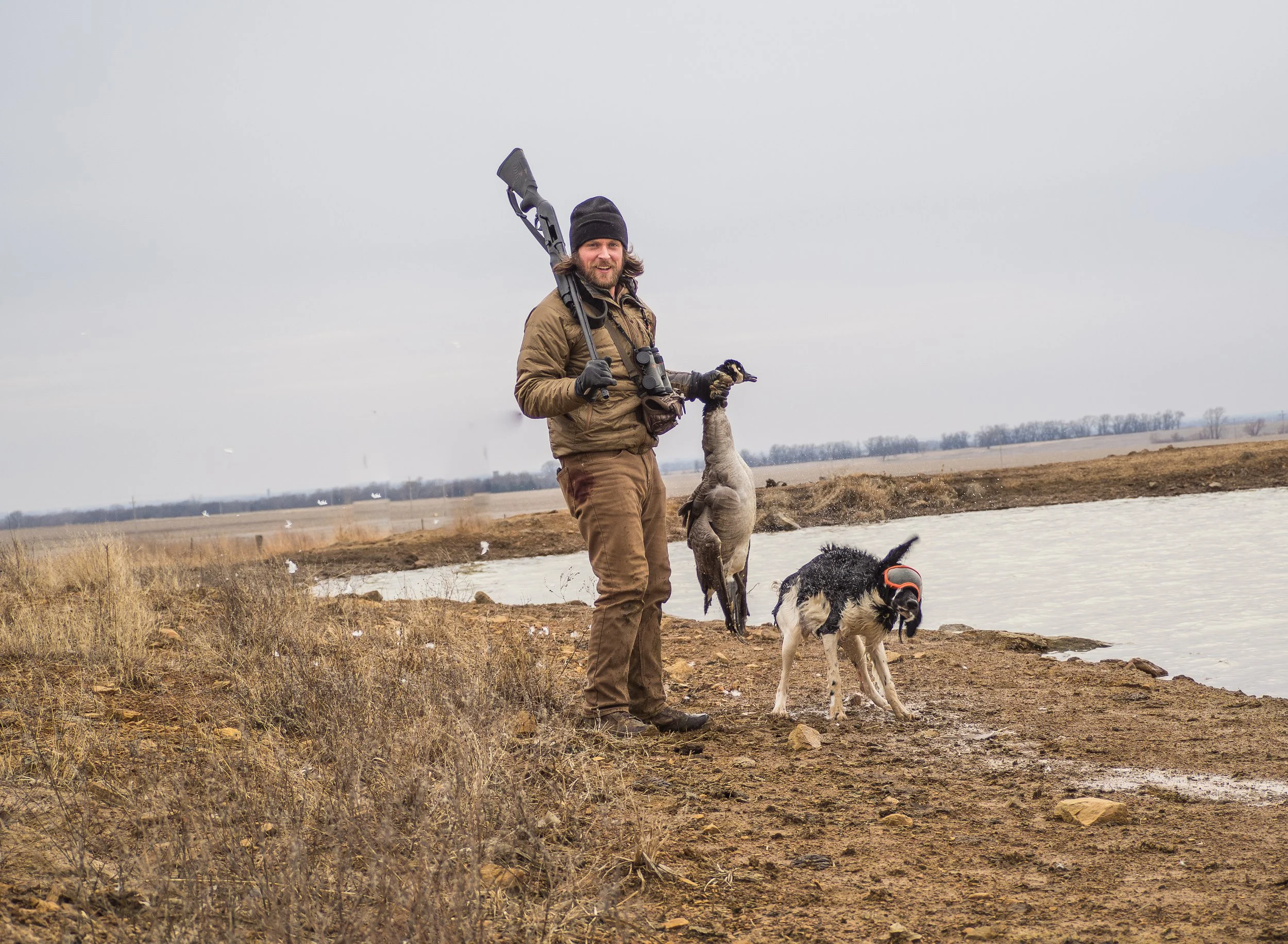 A man on a muddy shoreline holding a dead bird, with a hunting firearm over his shoulder, and two dogs nearby, in an outdoor setting with water and a cloudy sky.