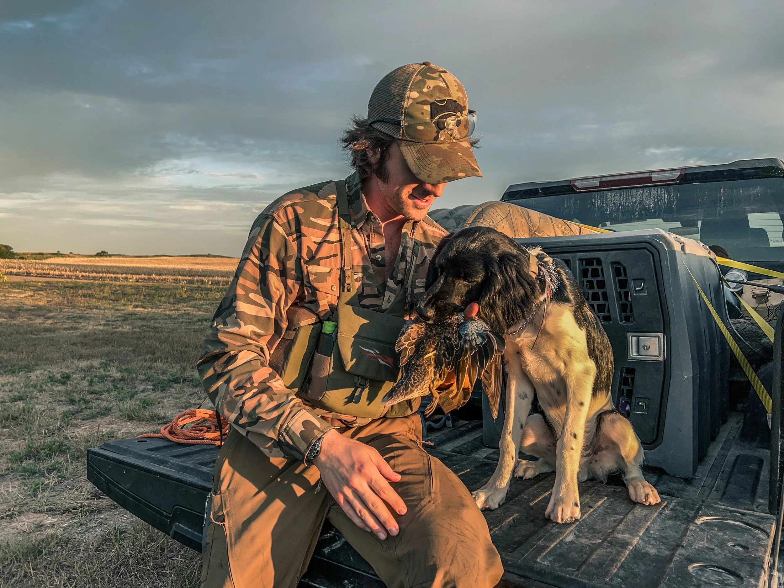 A man in camouflage clothing and cap sitting on a truck bed, holding a hunting dog with a bird in its mouth, in an open field during sunset.