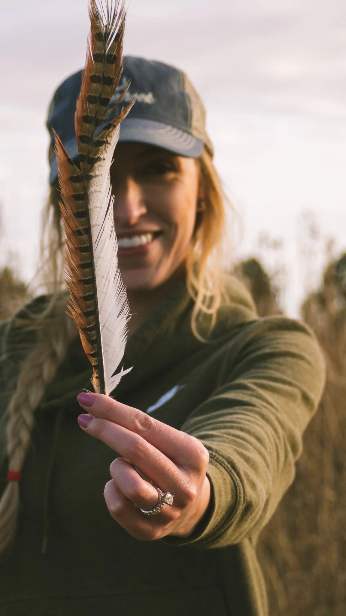 A woman smiling and holding a large pheasant tail feather close to the camera, outdoors in a natural setting with trees and a cloudy sky.