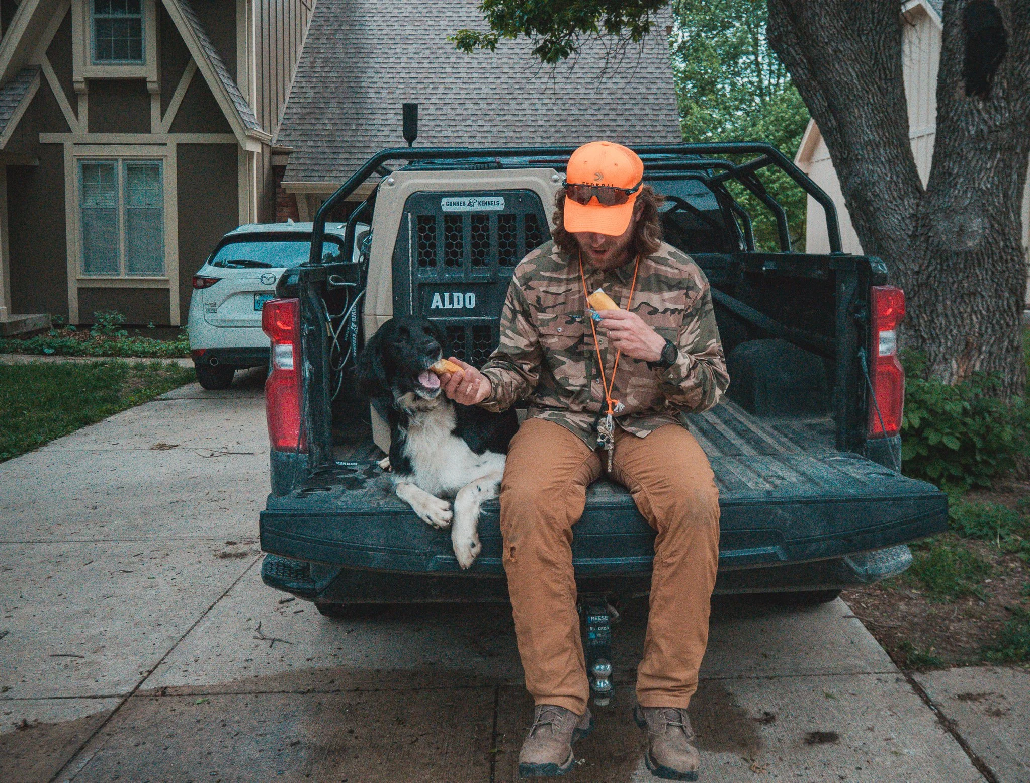A man in camouflage jacket, tan pants, orange cap, and sunglasses sitting on the tailgate of a black pickup truck, eating a popsicle with a black and white dog sitting beside him also holding a popsicle, in a residential neighborhood with cars and ho