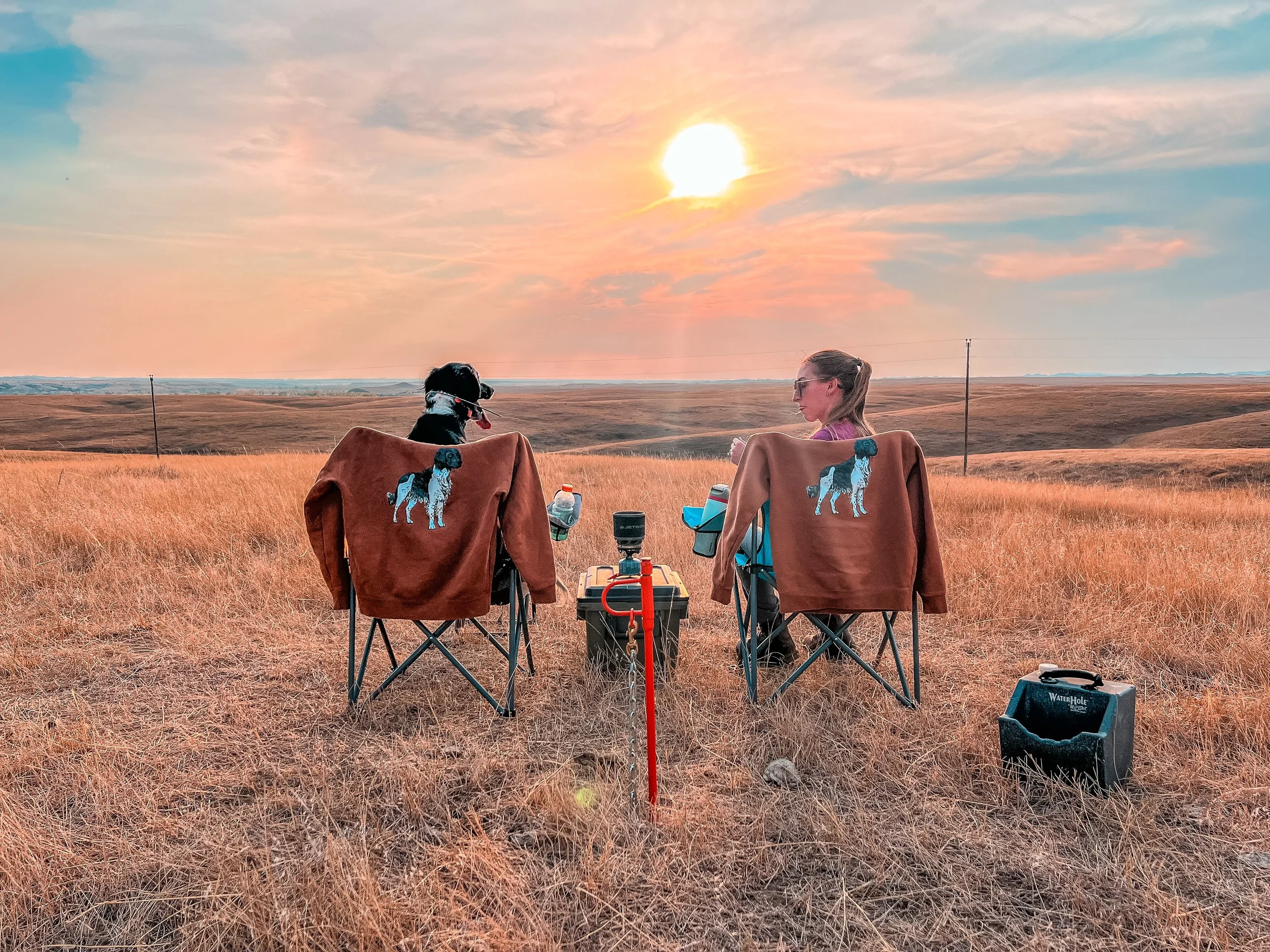 Two women and a dog sitting on camping chairs in a grassy field at sunset, overlooking a vast open landscape with a wire fence and power lines, each woman with a brown jacket featuring a dog logo on the back.