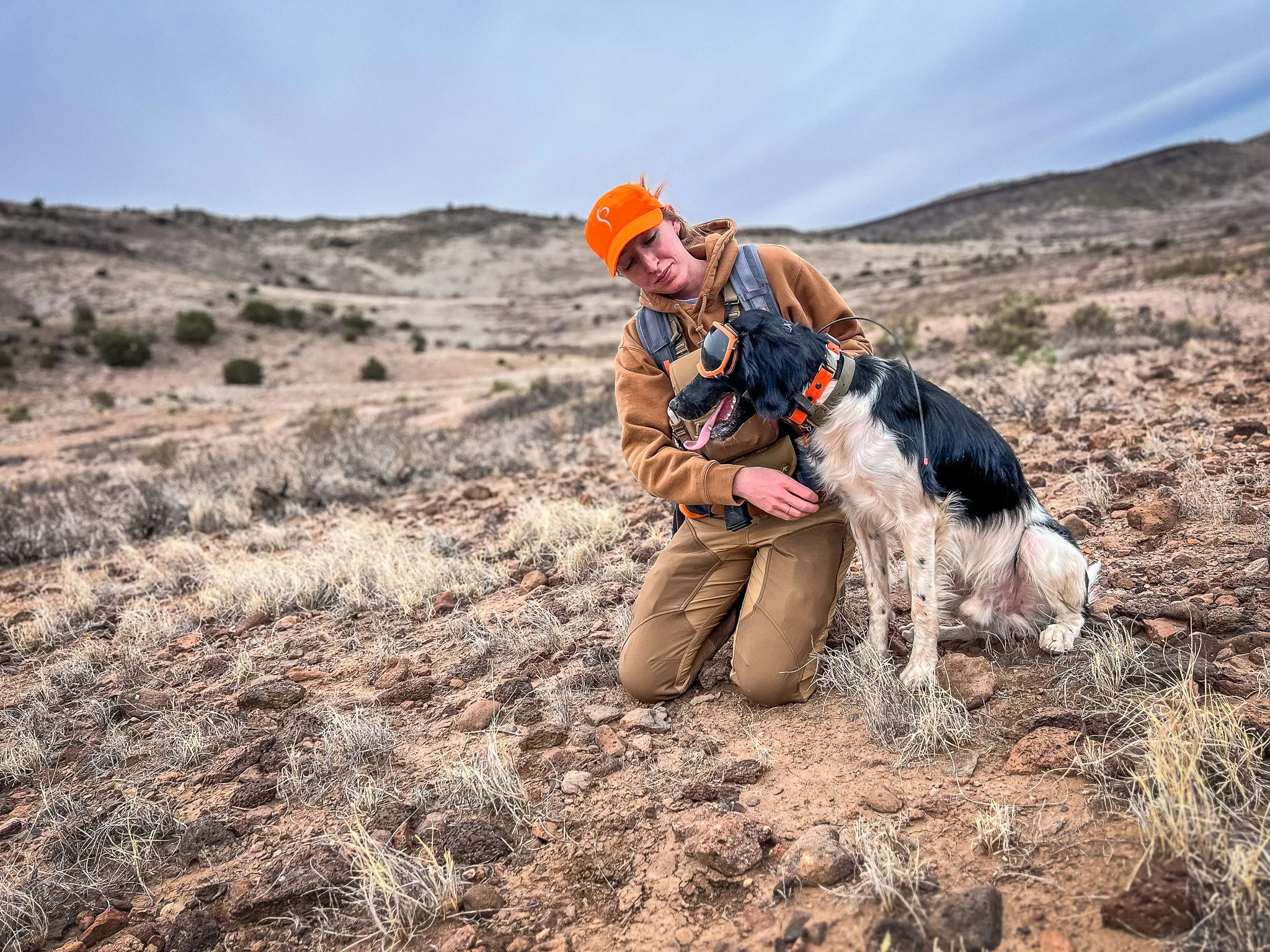 A woman kneeling in a desert landscape with rocky terrain and sparse vegetation, wearing a brown jacket, khaki pants, an orange helmet, and a backpack, holding a black and white dog wearing goggles and a collar.