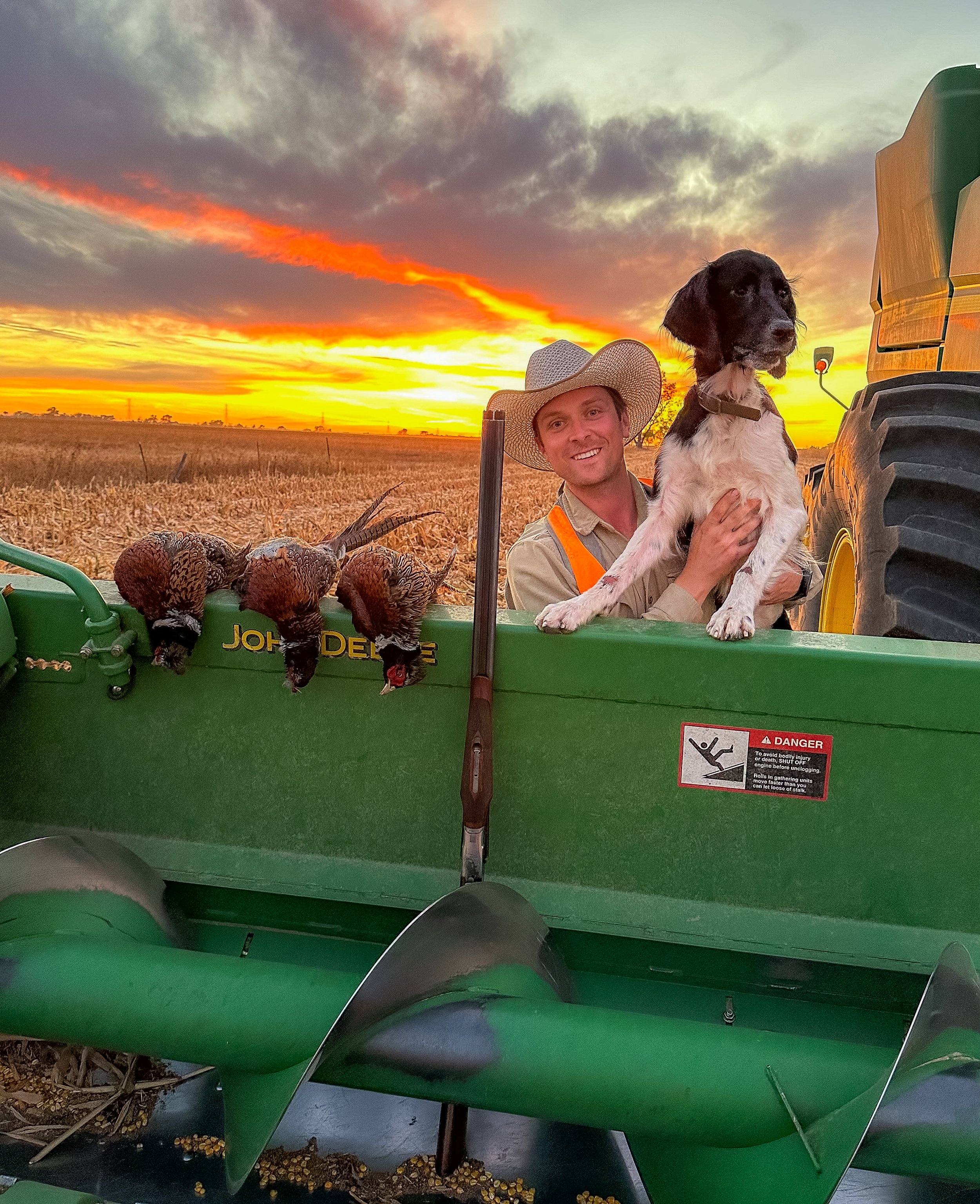 A man with a wide-brimmed hat and a dog in a tractor at sunset. Three dead birds are in the tractor bed, with a farm field in the background.