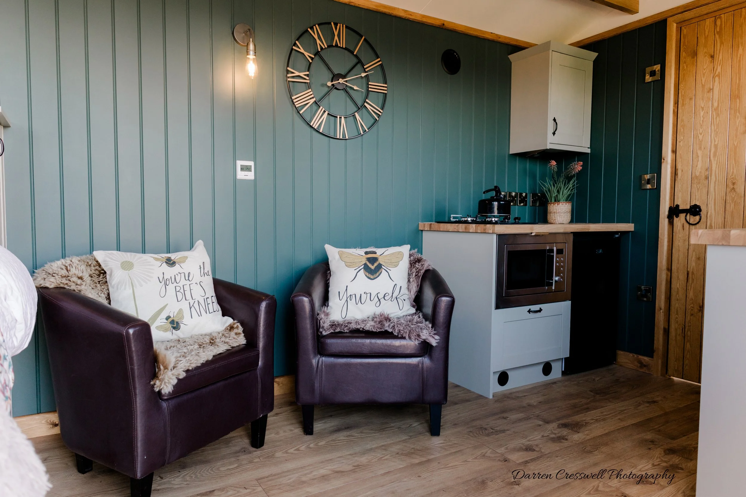 Living area with two brown leather armchairs with decorative pillows, one pillow reading 'You're the BEE'S Knees' and the other 'yourselF' with a bee illustration, against a green paneled wall. A large wall clock, a wall-mounted light with exposed bu