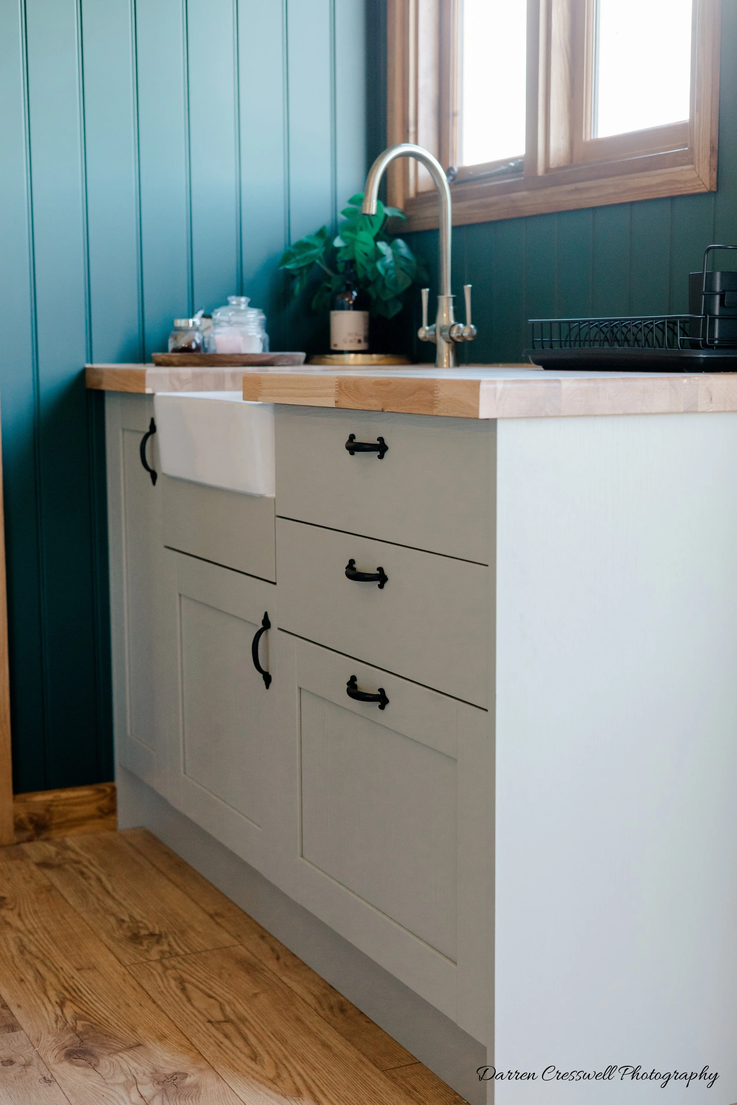 A kitchen with white cabinets, a wooden countertop, a farmhouse sink, a brass faucet, green wall paneling, a window, and a potted plant.