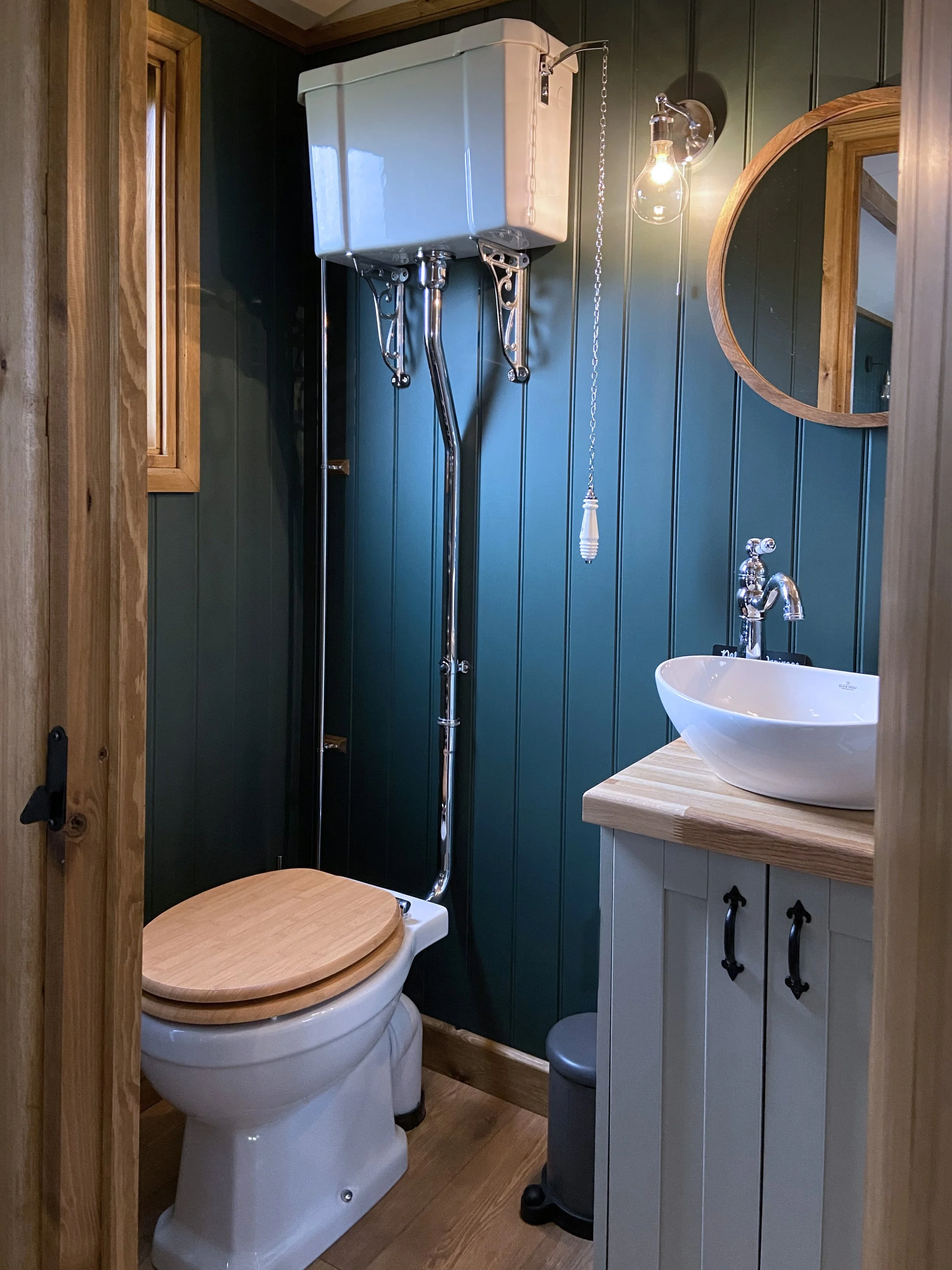 Small bathroom with dark green wooden paneling, a white toilet with a wooden seat, a small wooden cabinet with a vessel sink, a round mirror, and a wall light fixture with exposed bulbs.