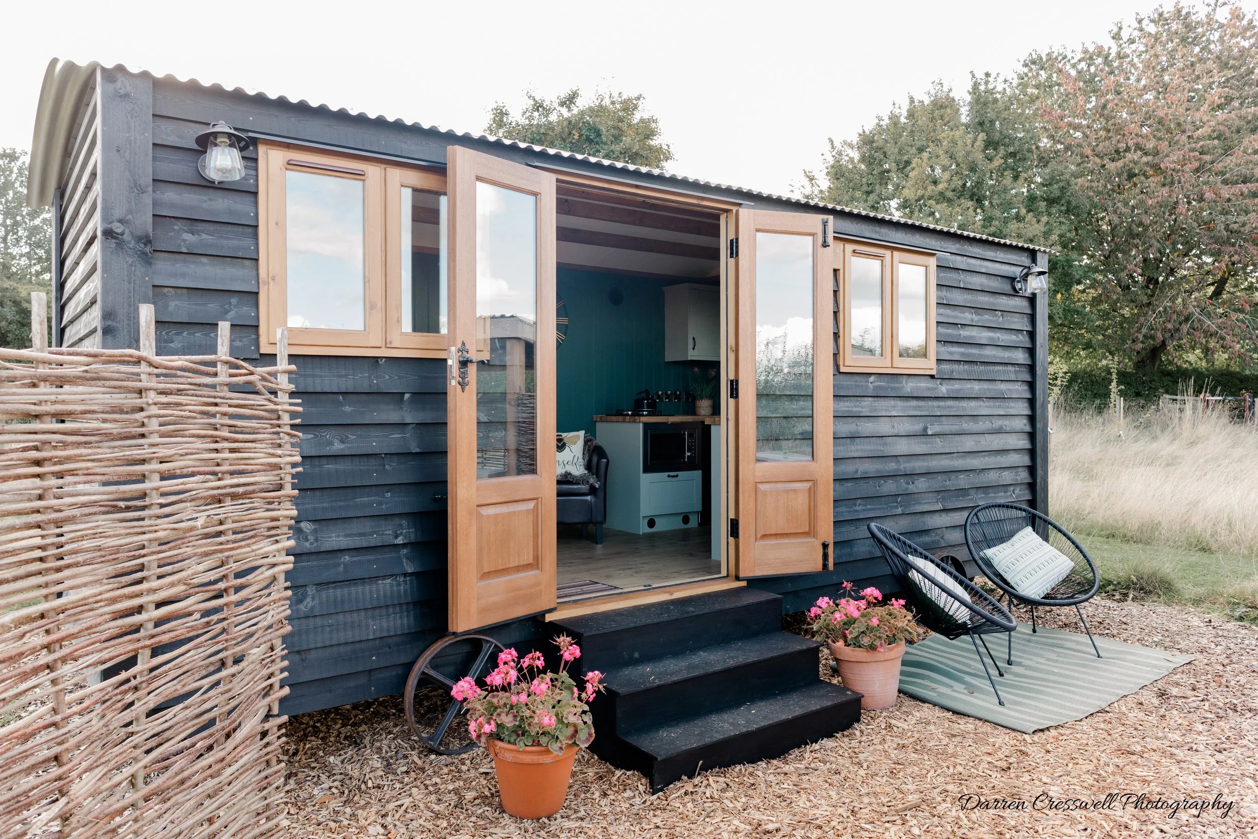 Small black wooden shepherds hut with open double glass doors leading to a cozy interior, surrounded by a grassy field with trees, decorated with potted flowers, outdoor chairs, and a woven fence.